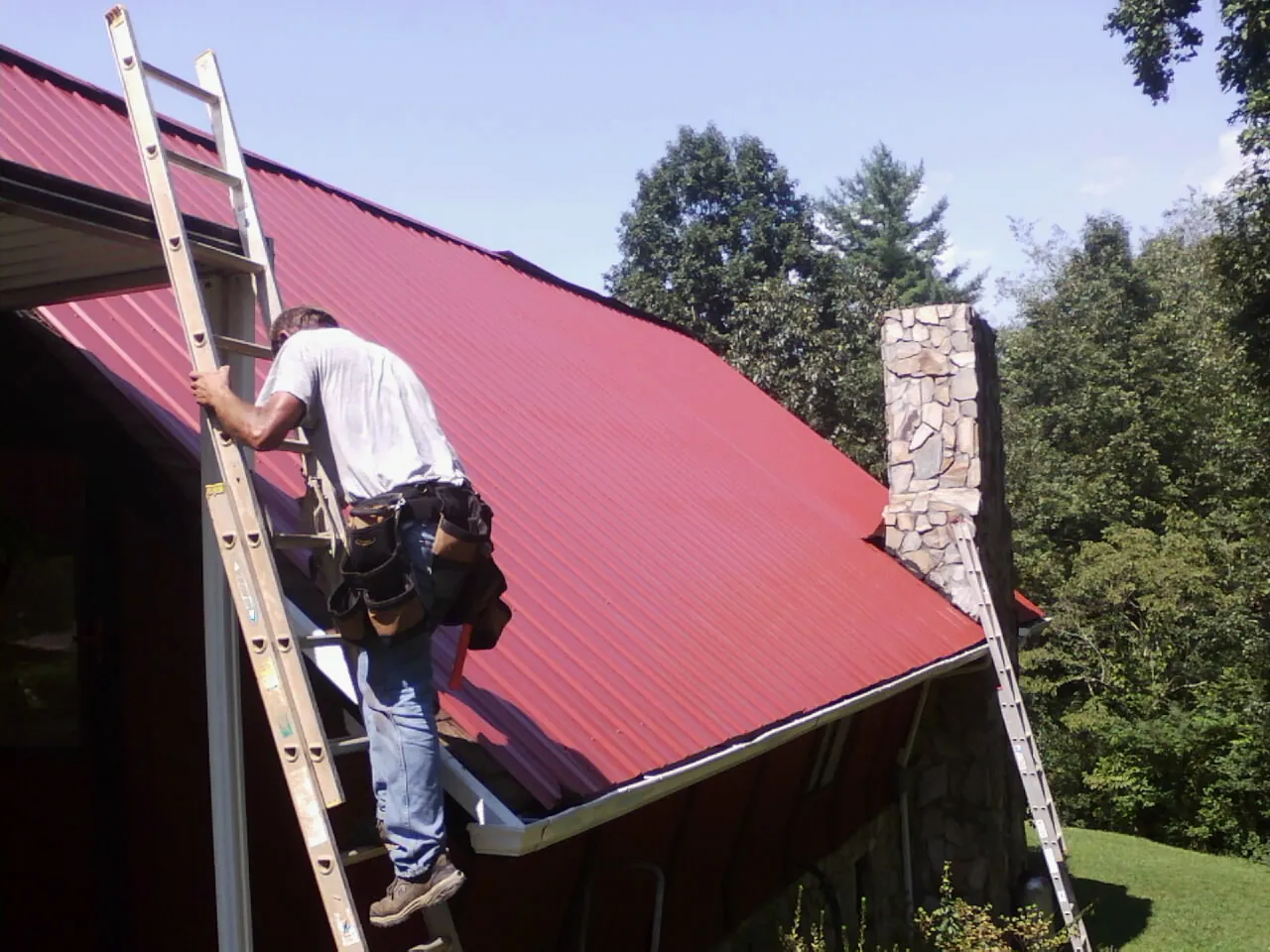 Roofer on a ladder beside a red metal roof and stone chimney — Soffit & Fascia Repair by Roofing Brothers in Pawhuska